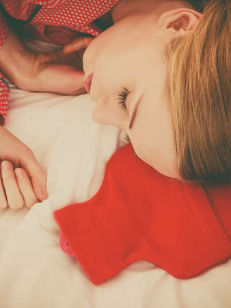Woman lying on bed sleeping with warm red hot water bottle. Fever, flu treatment objects concept.の写真素材