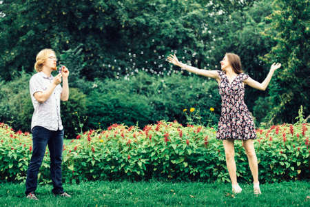 Happy funny hipster couple playing together blowing soap bubbles outdoor in spring park.