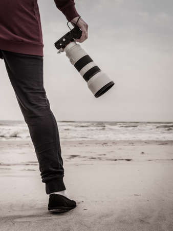 Unrecognizable woman having leisure time walking on beach during autumnal weather holding professional photography camera.の写真素材