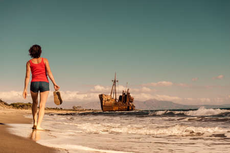 Travel freedom. Mature tourist woman walking on beach enjoying summer vacation. An old abandoned shipwreck, wrecked boat in the backgroundの写真素材