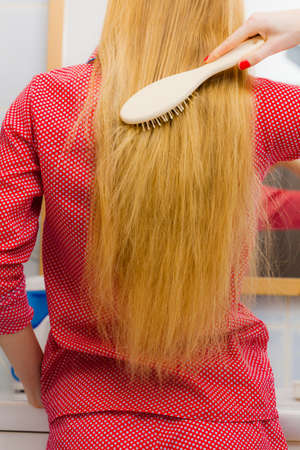 Young woman combing brushing her long blonde smooth hair in bathroom, using brush, looking in mirror. Girl taking care refreshing her hairstyle. Haircare concept.の写真素材