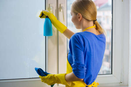 Young woman in yellow gloves cleaning window pane at home with rag and spray detergent. Cleaning...