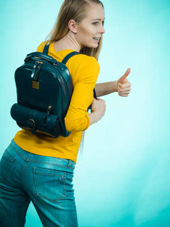 Happy young blonde teenage girl going to school or college wearing backpack showing thumbs up on blue.の写真素材