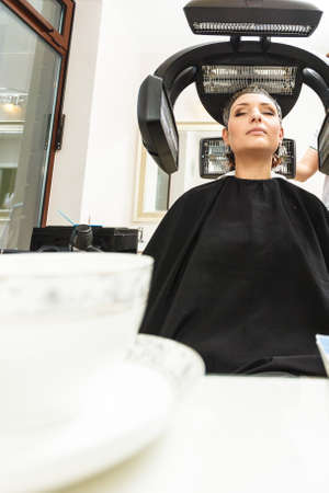 Haircare, relaxation and hairstyling concept. Woman sitting in black cape getting her hair dried under machine, cup of coffee in the foregroundの写真素材