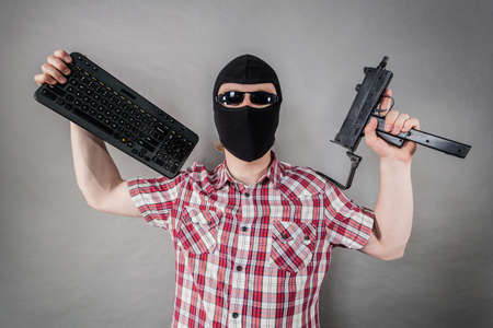 Crazy hacker man. Unrecognizable guy wearing black balaclava holding gun and keyboard. Hate speech on the internet.の写真素材