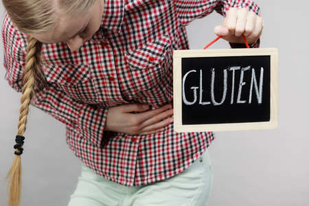 Wheat allergy problems concept. Woman having stomach cramps feeling very unwell holding board with gluten text sign written.の写真素材
