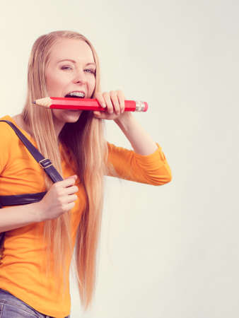 Happy young blonde teenage girl going to school or college wearing backpack and biting big pencil.の写真素材