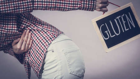 Wheat allergy problems concept. Woman having stomach cramps feeling very unwell holding board with gluten text sign written.の写真素材