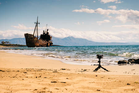 Camera on little tripod taking picture film video from coastline with rusty shipwreckの写真素材
