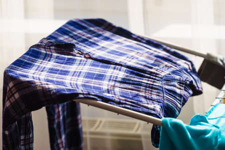 Many colorful clothes from laundry hanging on drying rack at home next to window.
