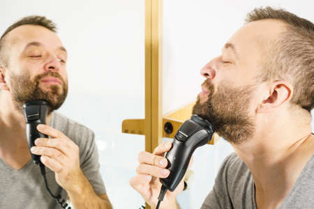 Bearded man looking at himself in mirror trimmng, shaving his beard using electric timmer razor.