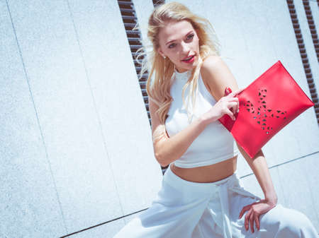 Glamorous female fashion model wearing casual elegant outfit white crop top. Blonde woman holding red handbag.. Summer urban style.の写真素材
