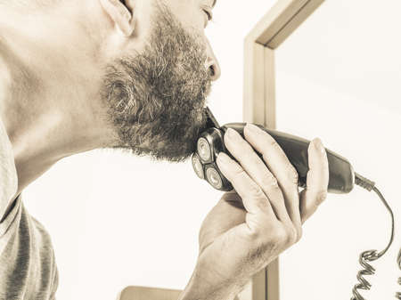 Bearded man looking at himself in mirror trimmng, shaving his beard using electric timmer razor.の写真素材
