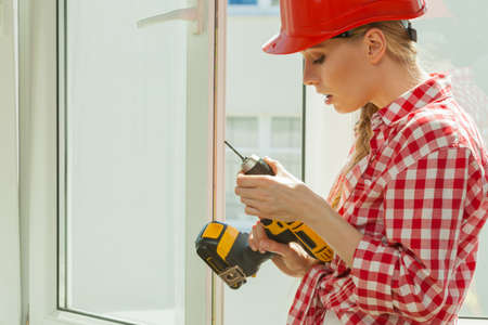 Young woman wearing helmet using power drill and tools for work at home, installing windows. Girl working at flat remodeling. Building, repair and renovation.の写真素材