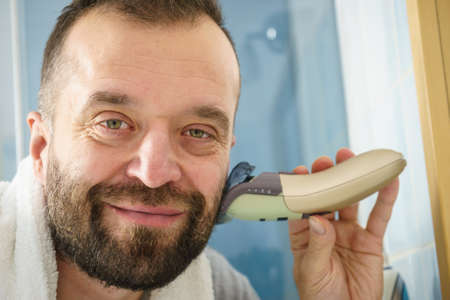 Bearded man looking at himself in mirror trimmng, shaving his beard using electric timmer razor.の写真素材