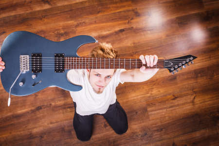 Young bearded man holding, raising his electric guitar. Hobby, music concept, on wooden floor. View from topの写真素材