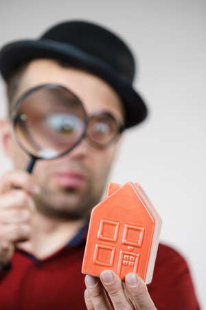 Funny looking adult man examining red model home using a magnifying glass, house inspection and real estate conceptの写真素材