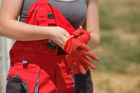 Woman in dungarees putting on red industrial safety worker gloves about to do work on construction site.の写真素材