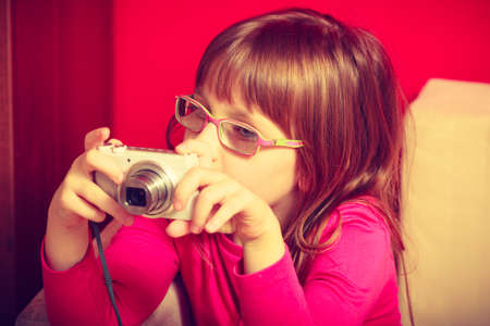 Childhood, childcare, discovering new hobbies and passion concept. Little toddler girl wearing eyeglasses sitting on sofa and playing with digital camera.の写真素材