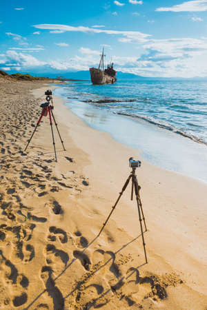 Professional camera on tripod taking picture film video from coastline with rusty shipwreckの写真素材