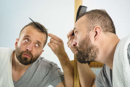 Adult man standing in front of the bathroom mirror brushing his short hair using comb. Guy investigating his receding hairlineの写真素材