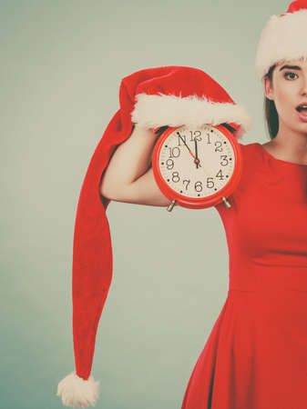 Xmas, seasonal clothing, christmas time concept. Worried woman wearing Santa Claus helper costume holding big red clock, waiting for celebrationの写真素材
