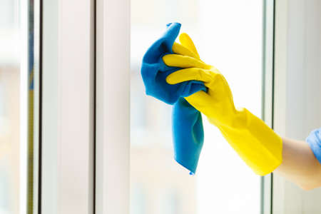 Female hand in yellow gloves cleaning window with blue rag and spray detergent. Spring cleanup,...