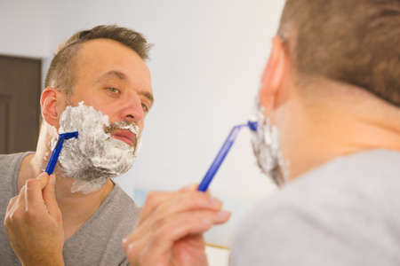Man with lots of shaving foam on his face standing in bathroom, looking at mirror, shaving his face...