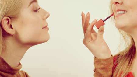 Make up artist friend applying another woman lipstick or lip gloss on lips. Two friends interested in visage.の写真素材
