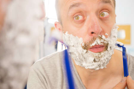 Man with lots of shaving foam on his face standing in bathroom, looking at mirror, shaving his face beard. Skincareの写真素材