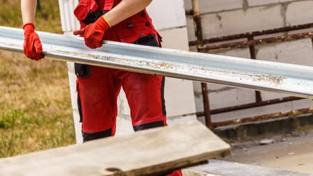Strong unrecognizable woman in dungarees carrying steel metal made gutter on her house construction...