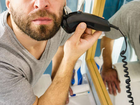 Bearded man looking at himself in mirror trimmng, shaving his beard using electric timmer razor.