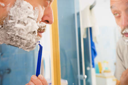 Man with lots of shaving foam on his face standing in bathroom, looking at mirror, shaving his face beard. Skincareの写真素材