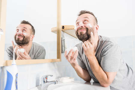 Happy funny adult guy applying moisturizer cream for male skin care. Man in front of bathroom mirror with anty aging lotion on face.の写真素材