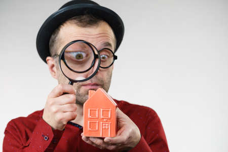 Funny looking adult man examining red model home using a magnifying glass, house inspection and real...