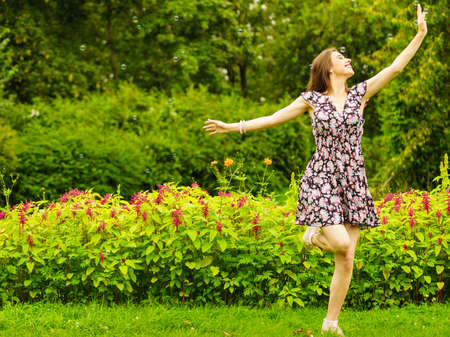 Pretty young adult woman wearing floral pattern dress dancing with soap bubbles in park during warm spring weather.の写真素材