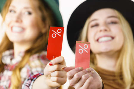 Female friends on shopping showing tag with sale percentage sign enjoying cheap clothing.