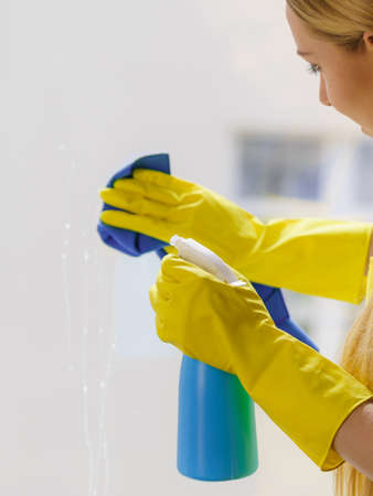 Young woman in yellow gloves cleaning window pane at home with rag and spray detergent. Cleaning...