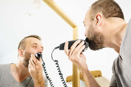 Bearded man looking at himself in mirror trimmng, shaving his beard using electric timmer razor.の写真素材