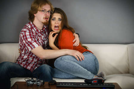 Funny couple sitting on sofa watching horror movie being scared hugging each other.