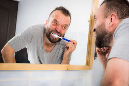 Adult man brushing his teeth looking at his bathroom mirror during morning hygiene routine.の写真素材