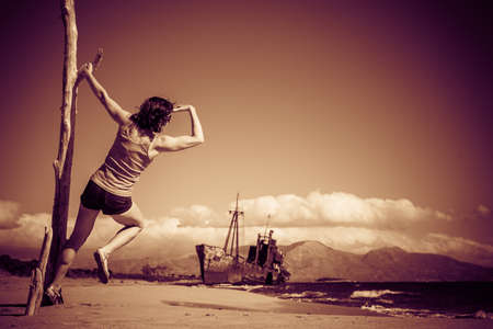 Travel freedom. Mature tourist woman on beach enjoying summer vacation. An old abandoned shipwreck, wrecked boat in the backgroundの写真素材