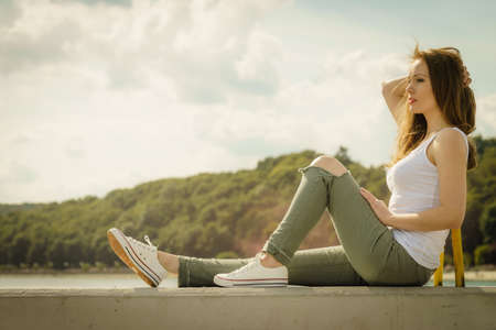 Young pretty fashion model woman sitting on concrete wall wearing white tank top and olive green trousers. Female walking outdoor during warm summer weather.の写真素材