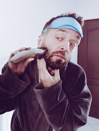Bearded man looking at himself in mirror trimmng, shaving his beard using electric timmer razor. Guy...
