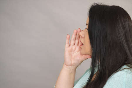 Profile view of adult woman whispering with her hand close to lips. Copy space on gray background in studio.の写真素材