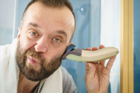 Bearded man looking at himself in mirror trimmng, shaving his beard using electric timmer razor.の写真素材