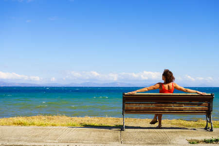 Travel freedom concept. Tourist mature woman sitting on bench on sea shore enjoying summer vacation, looking at oceanの写真素材