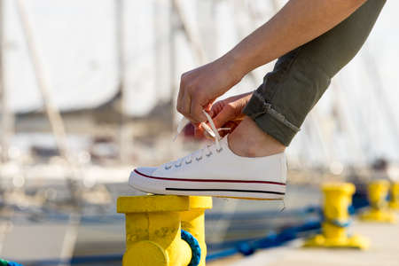 Person adjusting, tying up laces in white sneakers, casual shoes on yellow bollard pin. Outdoor footwear perfect for warm weather.の写真素材