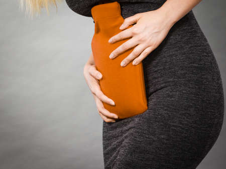 Unrecognizable woman having strong stomach ache. Female suffer on belly pain, holding hot red water...
