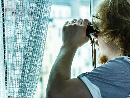 Young adult man spying at his neighbors, guy using binoculars looking through window.